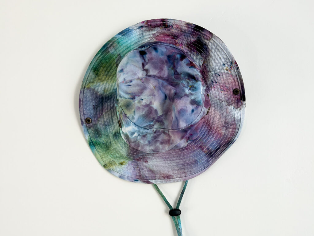 Hand-dyed bucket hat with ice dye technique in shades of blue, purple, and teal, hanging against a white wall