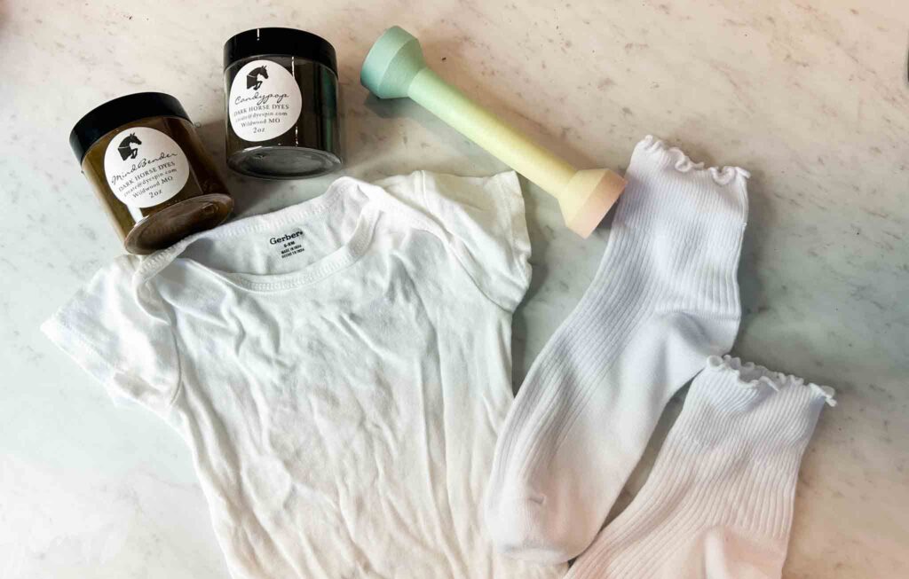 Tie dye supplies laid out including Dark Horse dyes, white Gerber onesie, socks, and tools