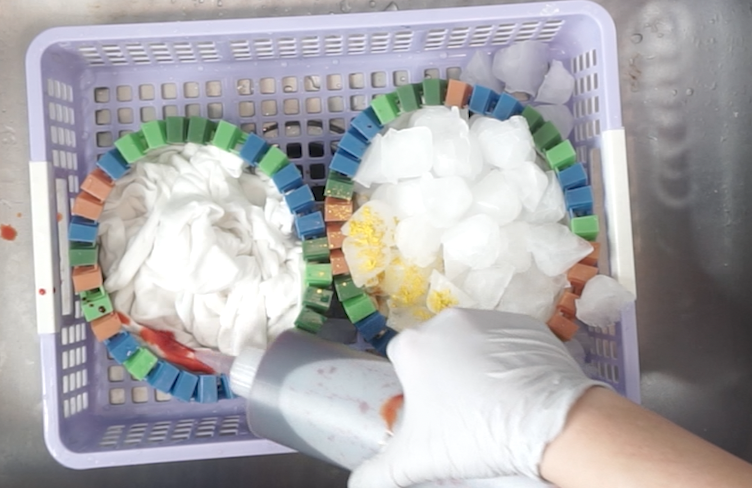 Side-by-side tie dye experiment with liquid dye and ice dye on scrunched white shirts in plastic baskets, one side with dye applied directly and the other covered in ice and powdered dye.