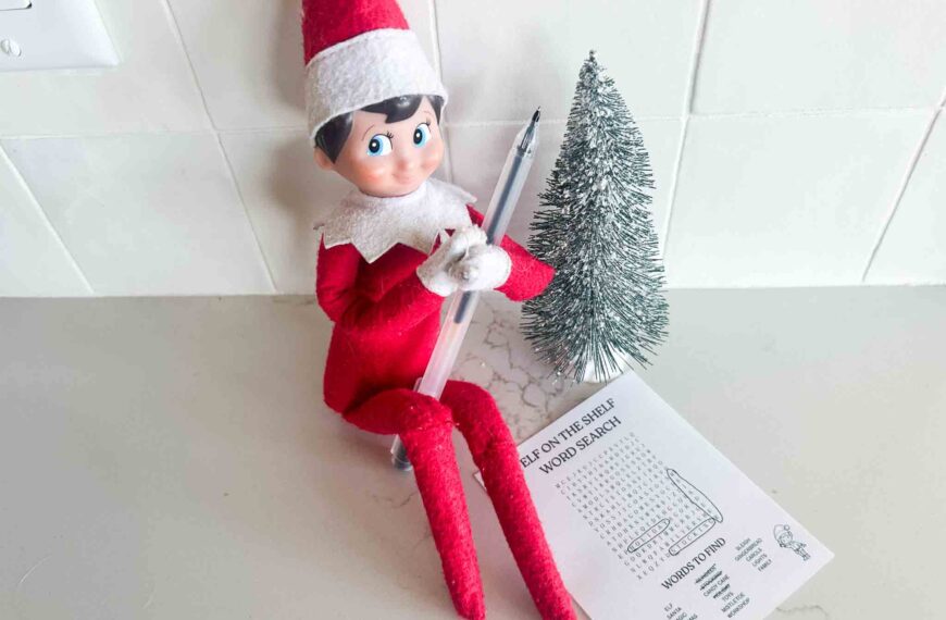 Elf on the Shelf sitting with a pen next to a Christmas word search printable and mini tree.
