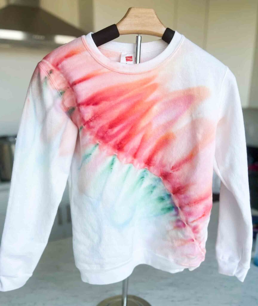 White sweatshirt with diagonal gravity fold tie dye design in soft red, orange, and green tones.