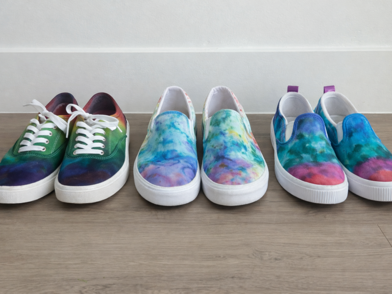 Tie dye shoes comparison showing three pairs of canvas shoes dyed using Sharpie, ice dye, and shaving cream techniques arranged side by side on a wooden floor.