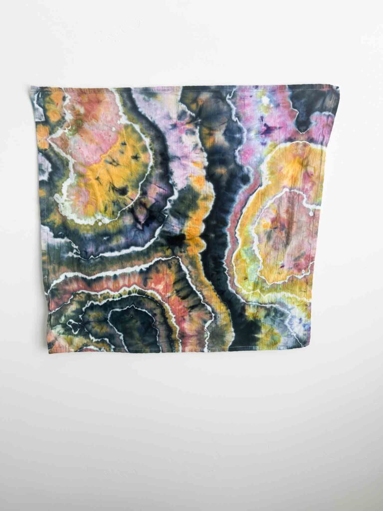 Square piece of geode ice dyed fabric in black, gold, orange, and pink tones displayed flat against a white wall