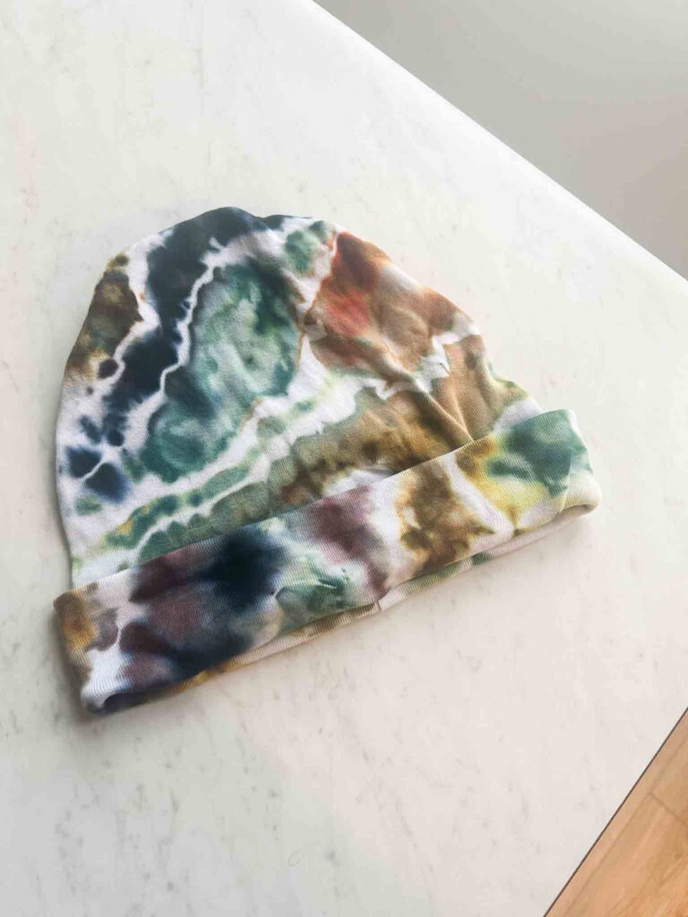 Ice dyed beanie hat in earthy green, teal, rust, and gold geode pattern laid flat on a white surface