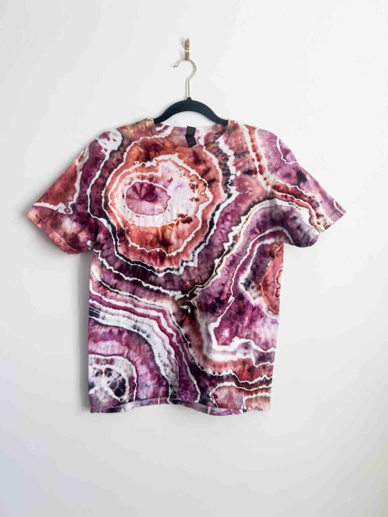 Geode ice dye t-shirt in shades of raspberry, mauve, rust, and white on a black hanger against a white wall