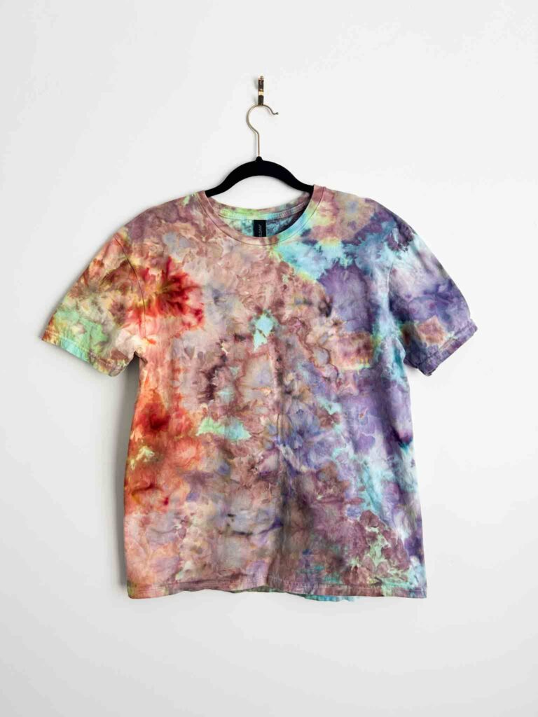 Front view of an ice dyed t-shirt in soft pink, purple, teal, and orange tones on a black hanger against a white wall