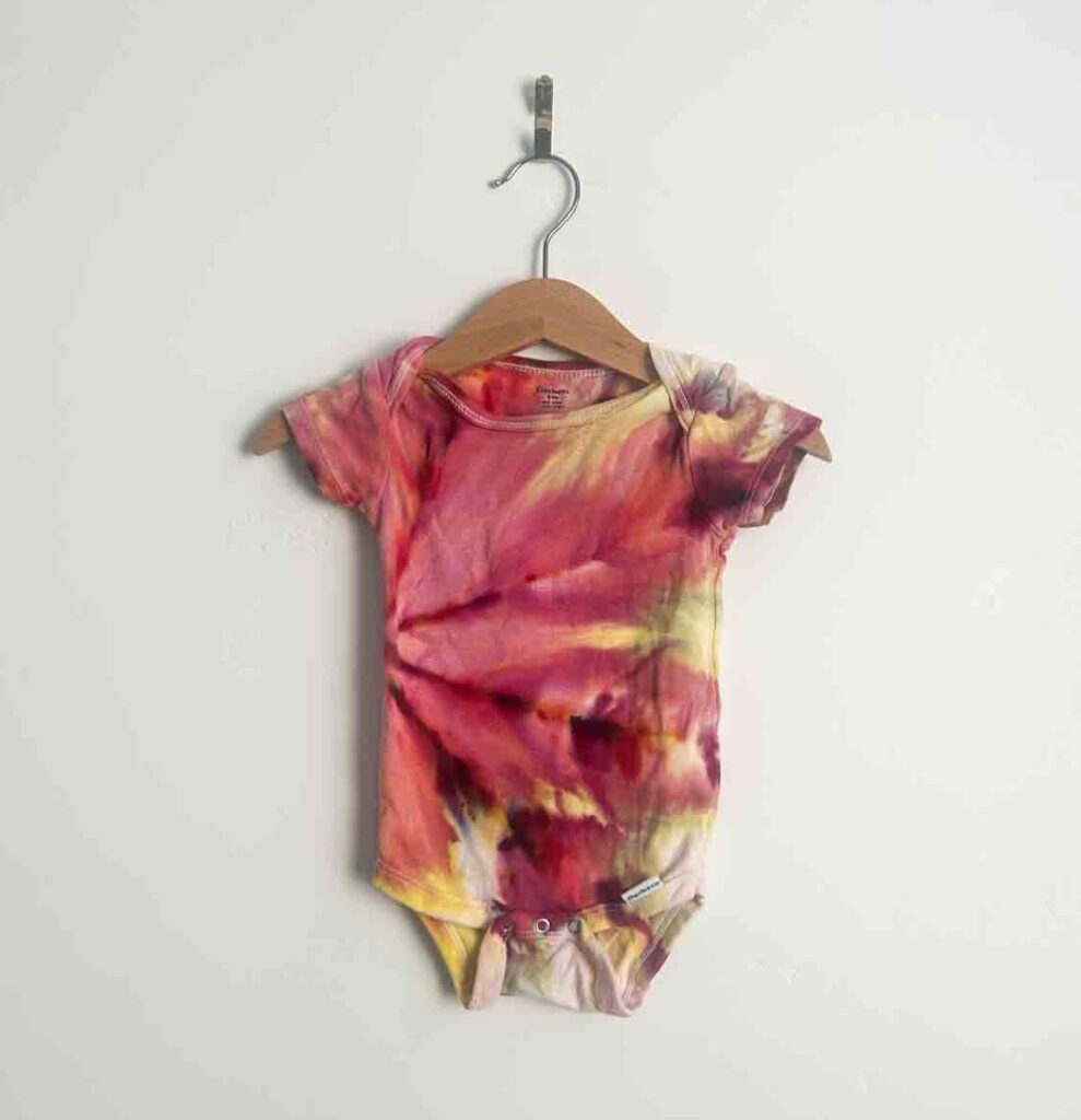 Tie dyed baby onesie in warm shades of pink, red, and yellow on a small wooden hanger against a white wall