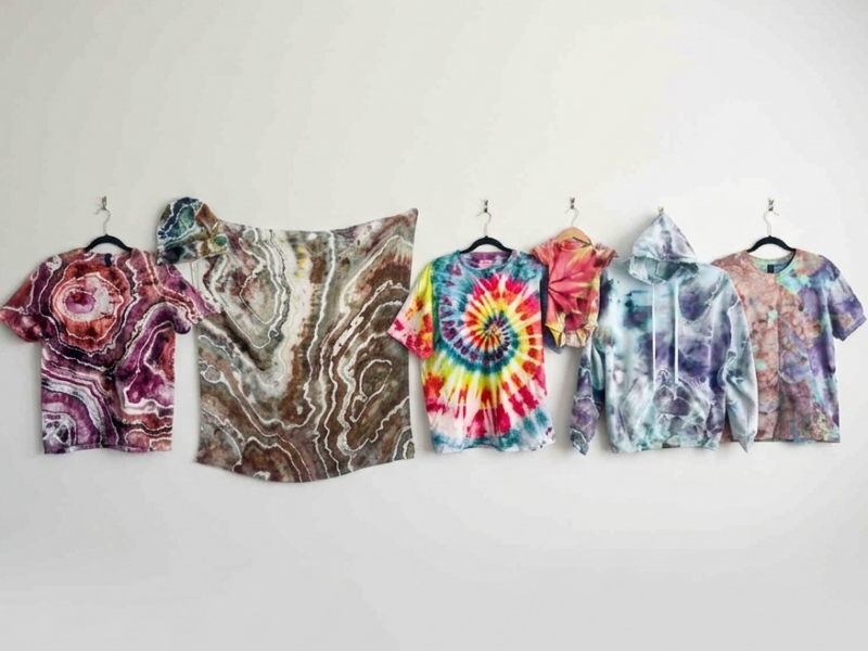 A collection of tie dyed and ice dyed pieces hung in a row against a white wall, including a geode t-shirt in pink and purple, a geode beanie and fabric panel in brown and gray, a rainbow spiral t-shirt, a pink and red onesie, a blue and purple ice dye hoodie, and a soft multicolor ice dye t-shirt