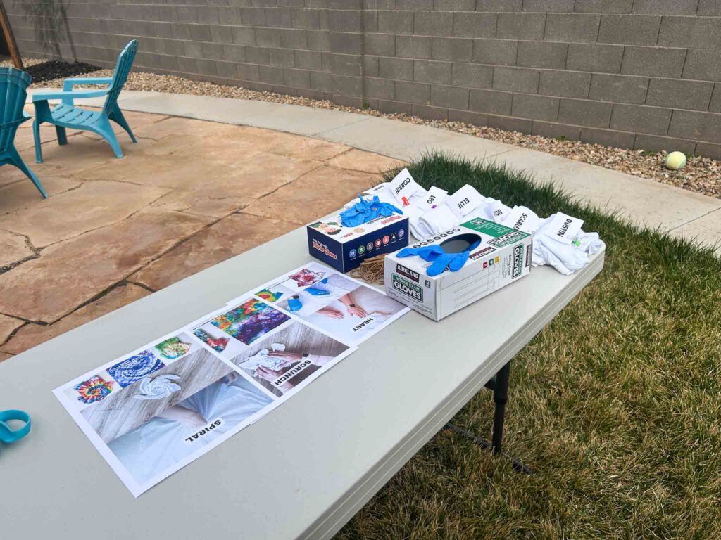 Tie dye party folding station with printed spiral, scrunch, and heart fold reference guides, nitrile glove boxes, rubber bands, and pre-labeled white shirts with guest names
