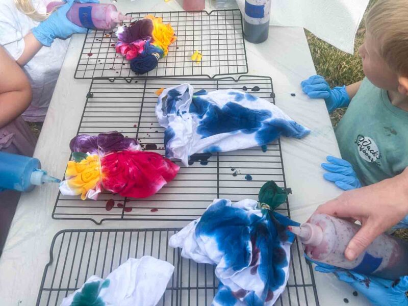 Several folded and banded tie dye shirts in progress on a cooling rack with children applying different colored dyes