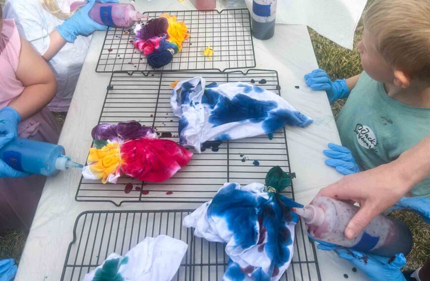 Several folded and banded tie dye shirts in progress on a cooling rack with children applying different colored dyes