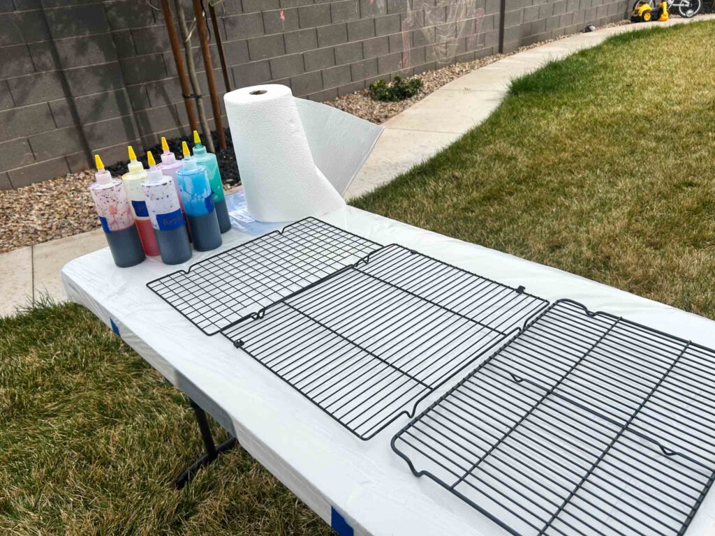 Outdoor tie dye party station with labeled squeeze bottles of dye, cooling racks on a plastic-covered folding table, and paper towels ready for guests