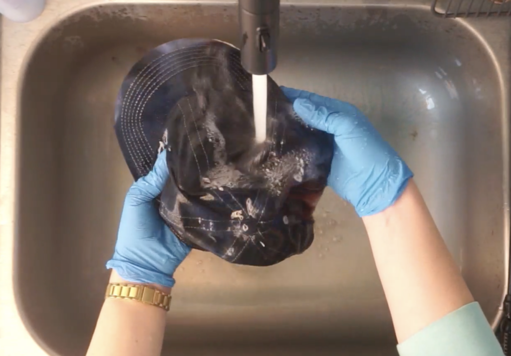 Rinsing a black ice dyed baseball hat under running water in a sink wearing blue gloves