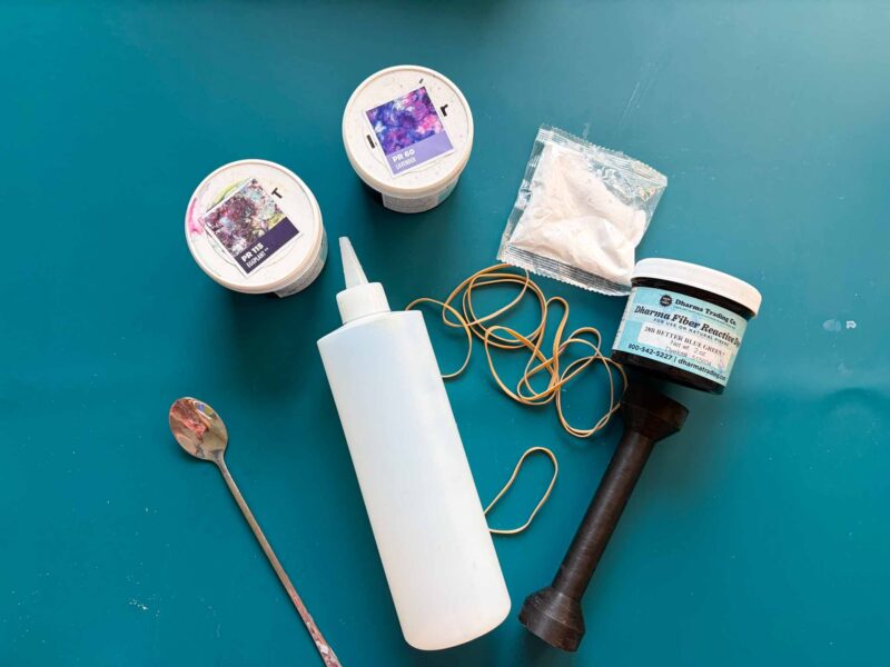 Tie dye supplies flat lay with Dharma fiber reactive dye, soda ash, squeeze bottle, rubber bands, and sinew puller